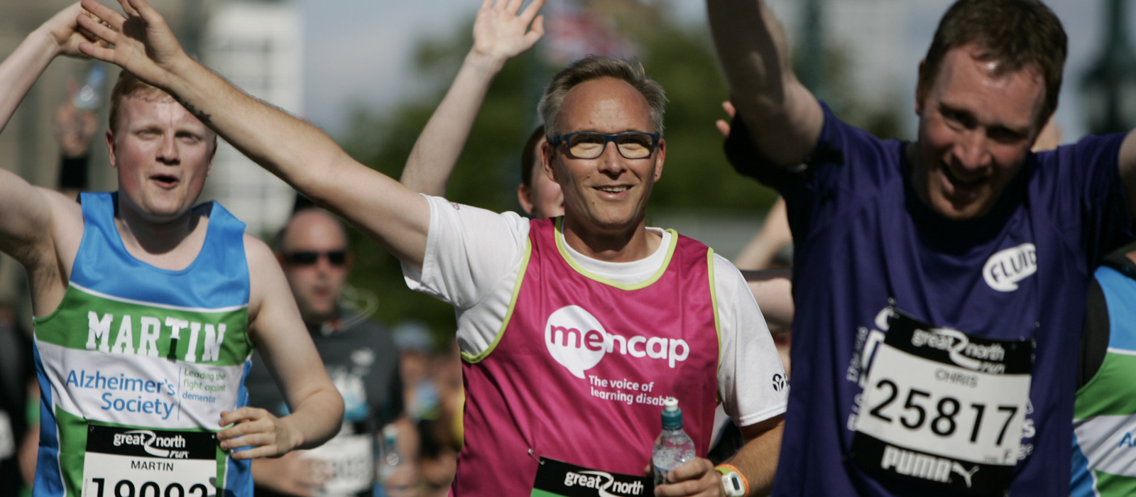 Group of runners taking part in outdoor running event on sunny day. A man in the middle of the photo has grey hair and glasses and is wearing a Mencap running vest whilst waving his arm in the air.