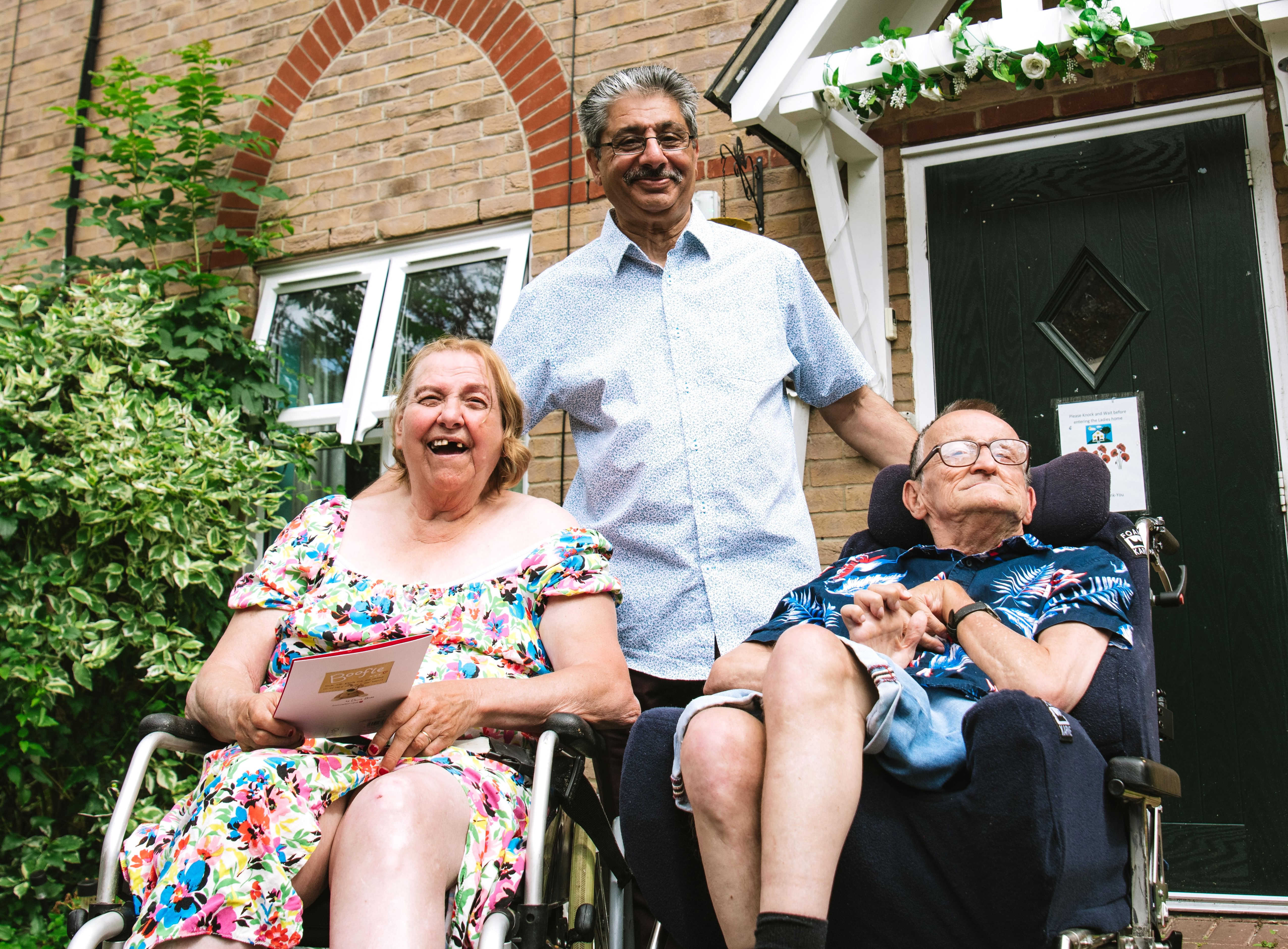 A friendly older man stands behind an older couple both in wheelchairs. They are in front of a black door & there is a bush to the left hand side