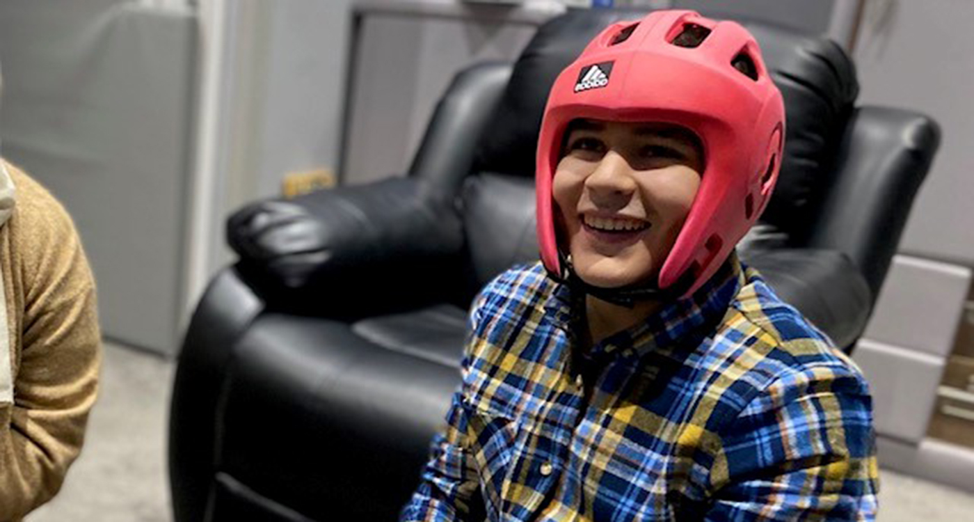 Young man smiling whilst sat on the floor in living room He is wearing a blue and yellow chequered shirt and a pink protective hat.