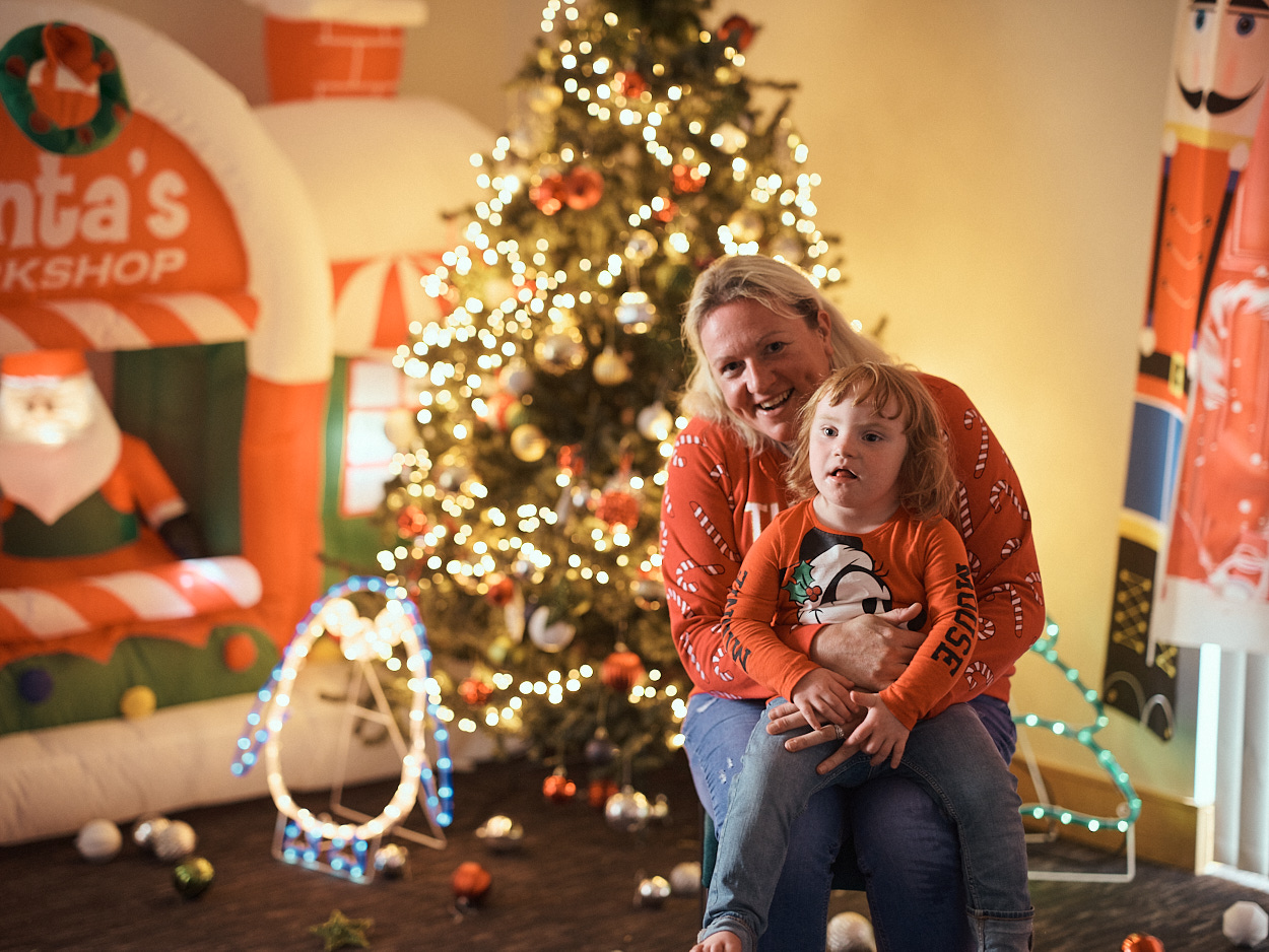 Lisa, sitting in front of a decorated Christmas tree, smiling warmly while holding her daughter Dakotah on her lap. They are both dressed in festive clothing, and there are Christmas decorations, including an inflatable Santa's Workshop.