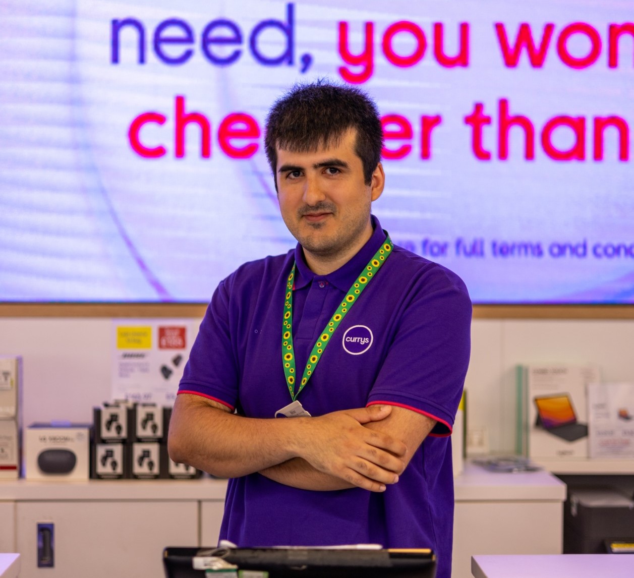 Niaz is pictured smiling proudly, arms folded, behind a counter in the Currys where he works. 