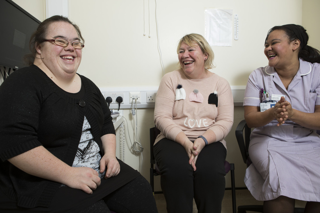 Three women, one of whom is a nurse, sat on chairs in nurses room.They are all laughing and smiling together.