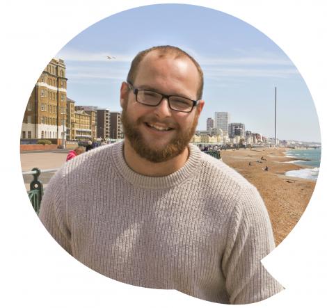 Young man wearing glasses stood on beach smiling into camera.