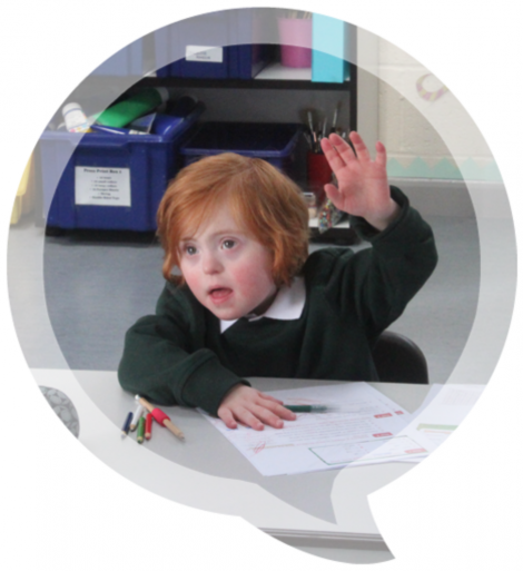 Young boy with red hair sat at table in classroom. He is raising his left hand in the air.