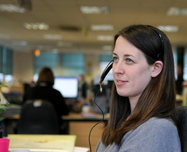 Woman with long brown hair sat at desk in an office wearing a telephone headset.