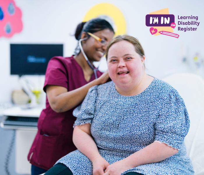 A woman with a learning disability wearing a blue dress smiles to the camera as a nurse does a health check.