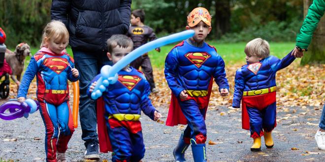 Four young children dressed in superman costumes walking in park.