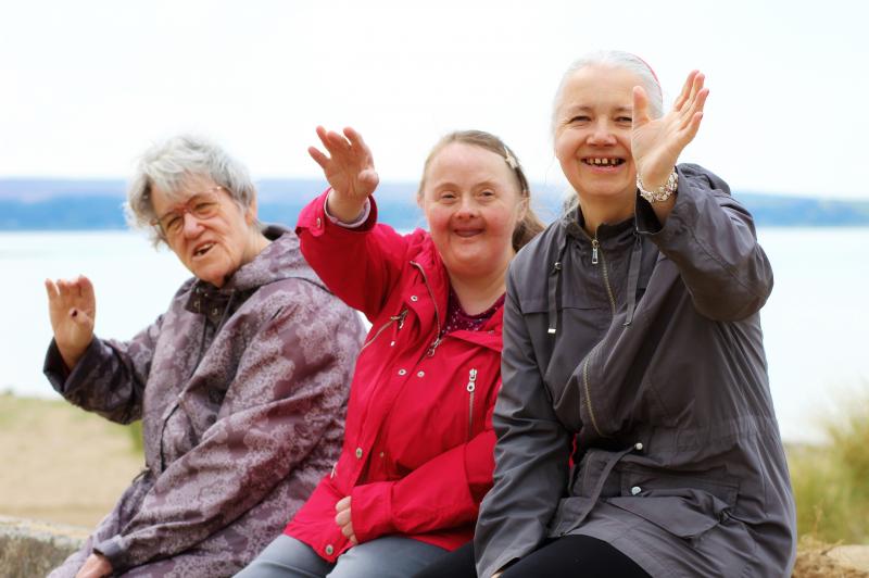 3 people with a learning disability sat on a coastal wall smiling and waving with the beach in the background